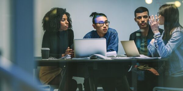 A diverse group of four people sit at a table with laptops and notepads, engaged in a focused discussion, conveying collaboration and concentration.